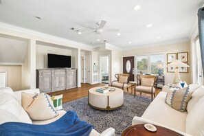 A spacious living room with a flat-screen TV, a round coffee table, and a ceiling fan.