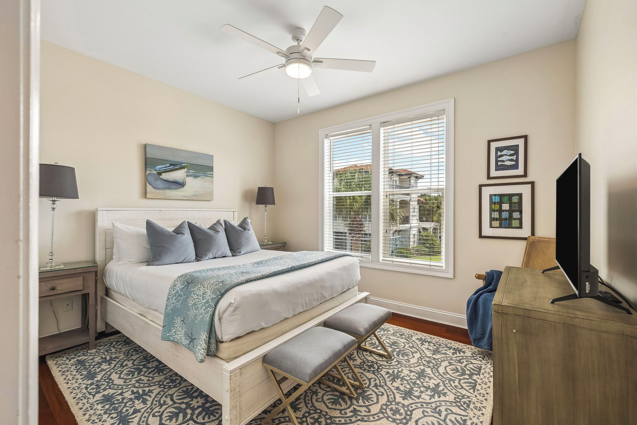 A bedroom with a bed, bedside lamps, a ceiling fan, a window with blinds, and artwork on the wall.