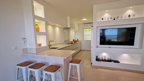 A modern kitchen with a central island, bar stools, a built-in TV, and a window with blinds.