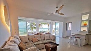 A modern living room with a sofa, coffee table, and a ceiling fan. Large windows offer a view of the ocean and palm trees.