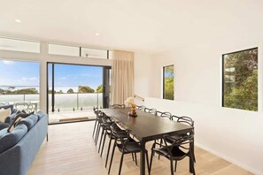 A modern dining area with a large table and chairs, large windows with a view, and a sofa in the foreground.