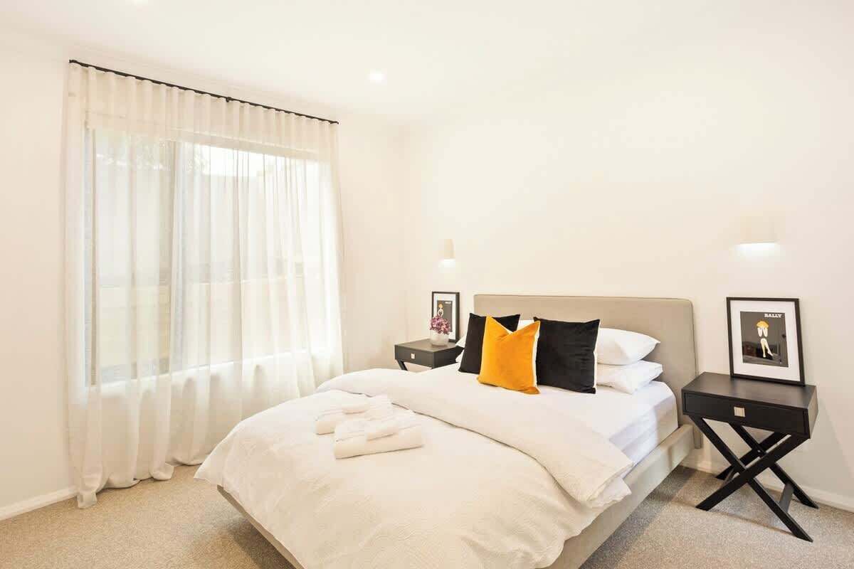 A hotel room with a bed, white curtains, a nightstand with a lamp, and a framed picture.