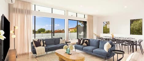A spacious living area with two sofas, a coffee table, and a dining set. Large windows provide a view of the outdoors.