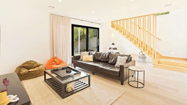 Una sala de estar moderna con un sofá, una mesa de centro y un sillón informal. Se ve una escalera con barandillas de madera al fondo.