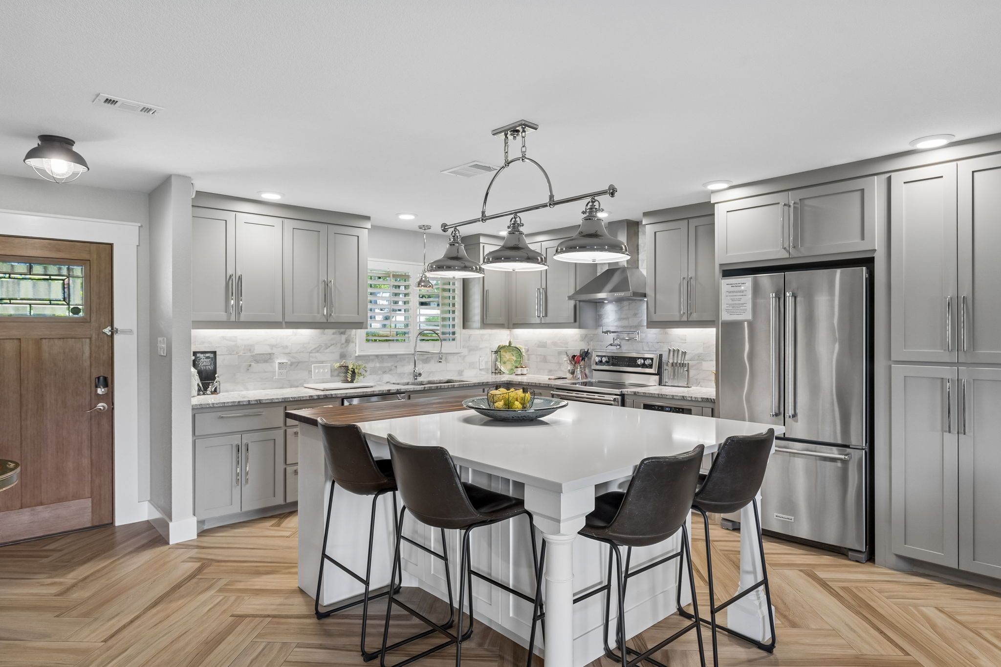 A modern kitchen with grey cabinets, a central island with bar stools, stainless steel appliances, and pendant lighting.