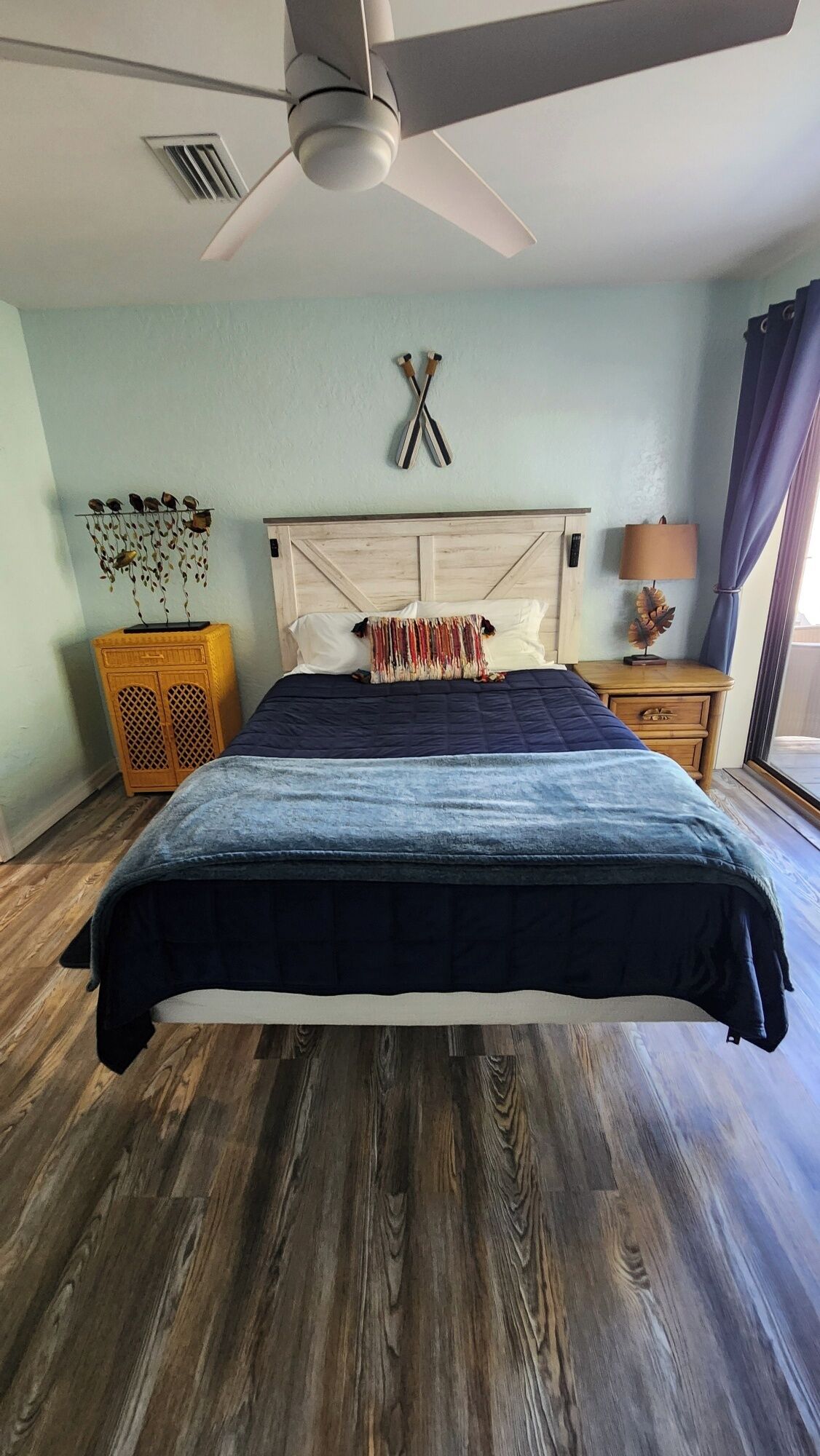 A bedroom with a bed, wooden floor, ceiling fan, wall-mounted decorations, and a window with curtains.