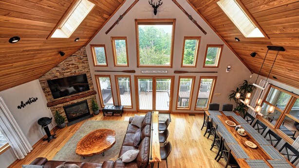 Un loft spacieux avec de hauts plafonds, des poutres en bois, une cheminée, un grand téléviseur, un coin repas et de nombreuses fenêtres offrant une vue sur l’extérieur.