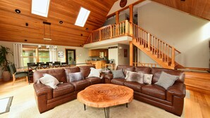 Un vaste salon comprenant un grand canapé d’angle en cuir marron, une table basse ronde en bois et un escalier en bois menant à l’étage supérieur.