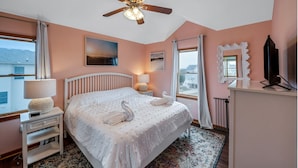 A bedroom with a bed, a ceiling fan, a bedside table with a lamp, a dresser, a mirror, and a window with curtains.