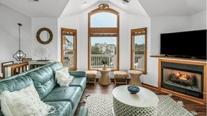 A living room with a teal sofa, a fireplace, a television, and a large window overlooking a house.
