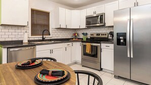 A modern kitchen with white cabinets, a stainless steel refrigerator, a microwave, and an oven. There is a wooden dining table with a colorful placemat.
