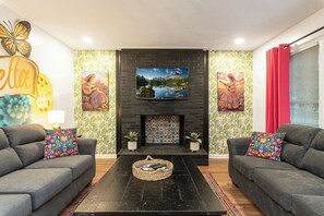 Una sala de estar con dos sofás grises, una mesa de centro negra, una chimenea con revestimiento de azulejos y un televisor de pantalla plana montado sobre la chimenea.
