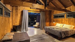 A log cabin interior with a loft bed, a sofa, a small cabinet, and a glass door leading to a balcony. - WoodStone Hotel Fazenda (Gramado)