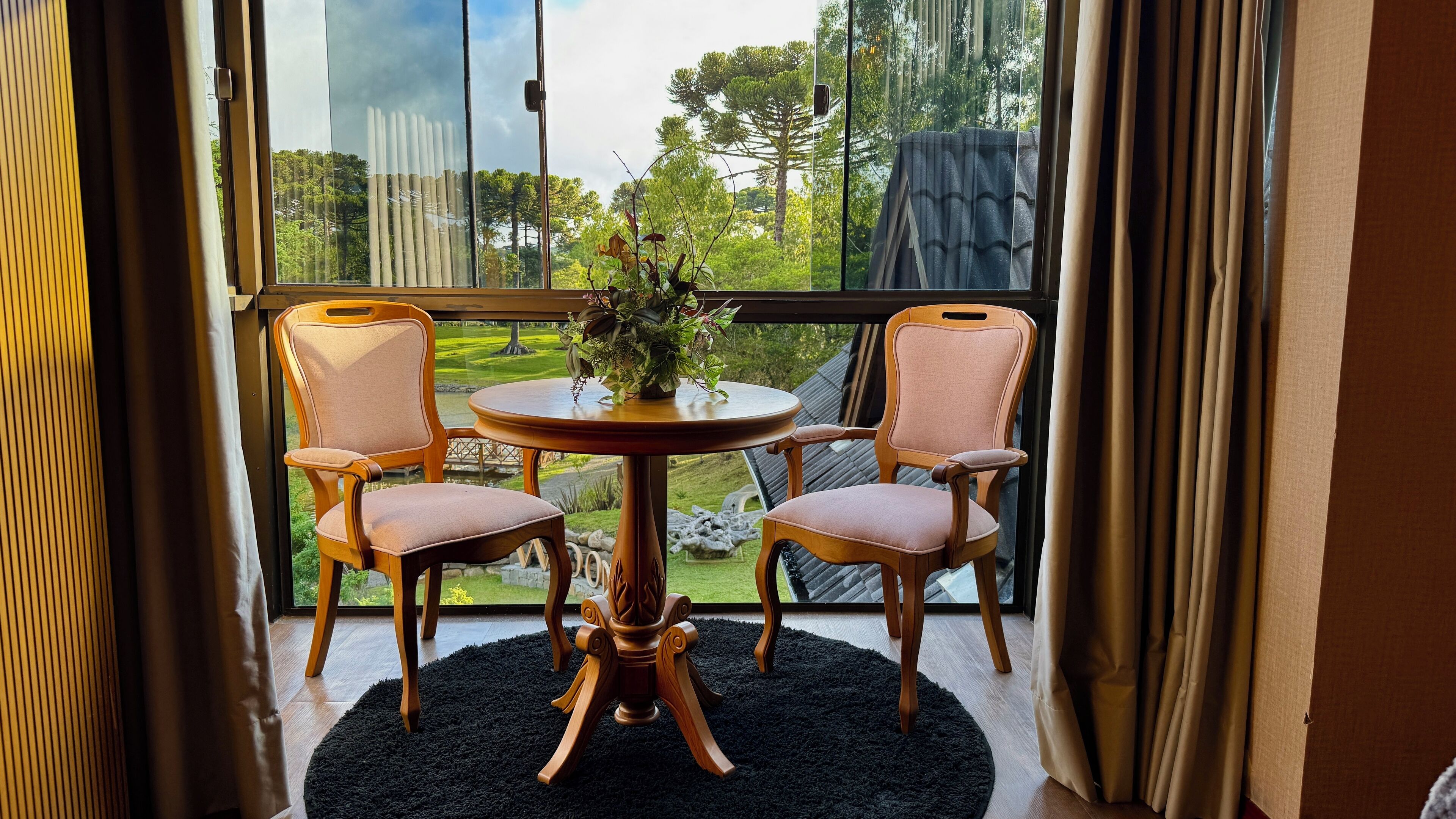A room with a large window, two wooden chairs, a round table, and a black rug.
