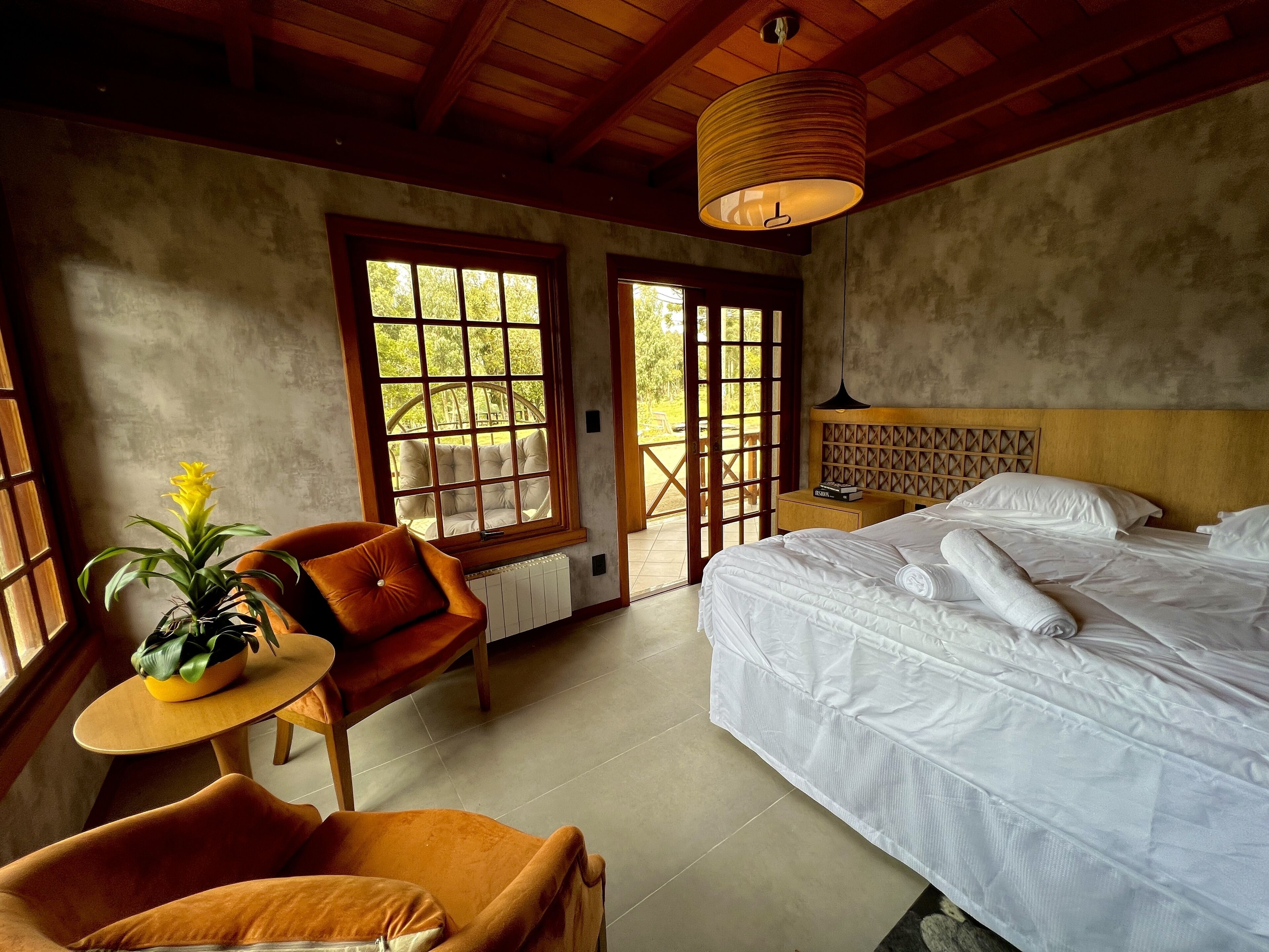 A bedroom with a large bed, a wooden headboard, a window with a view, a small table with a plant, and a chair.