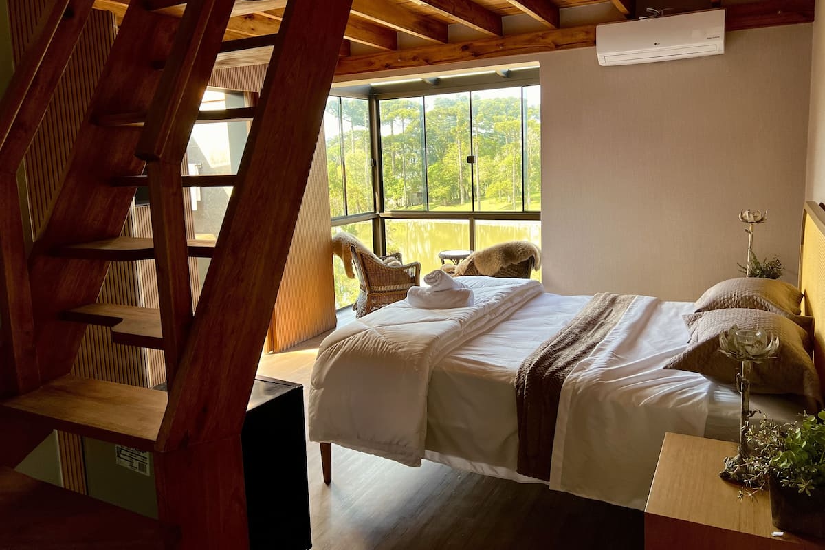 A bedroom with a bed, a wooden ladder, a window with a view of trees, and a wall-mounted air conditioner.