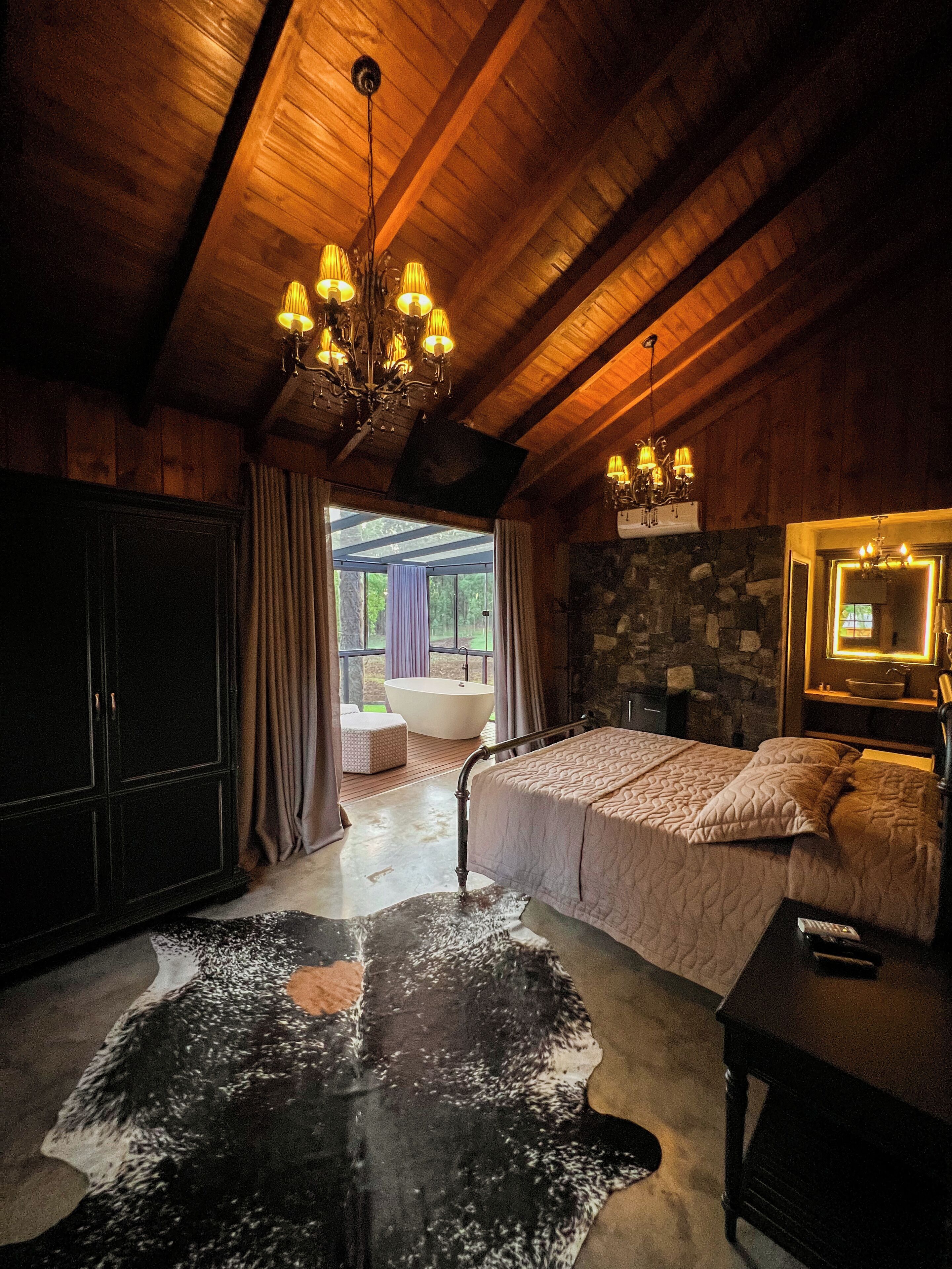 A bedroom with a large bed, a stone wall, a chandelier, and a view of the outdoors.