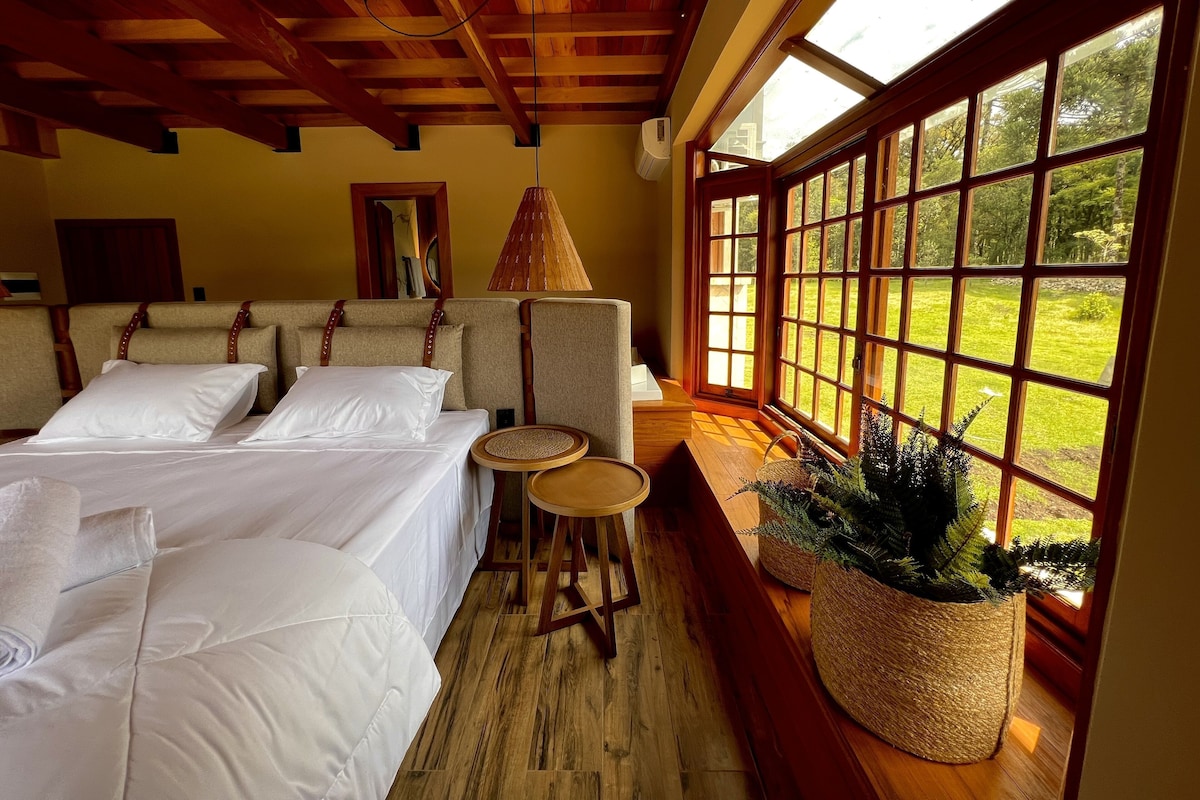 A room with a bed, a small table, a lamp, and large windows with a view of greenery.