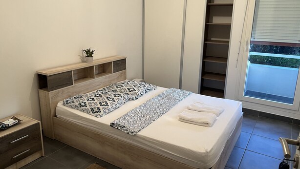 A neatly made bed with patterned bedding, a bedside table with a remote control, a small potted plant, and a view of a balcony through a window. - Lovely homestay hotel room, 10 minutes from the beach! (Cayenne)