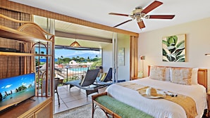 A hotel room with a bed, a TV, a balcony with a view of the beach, and a ceiling fan.