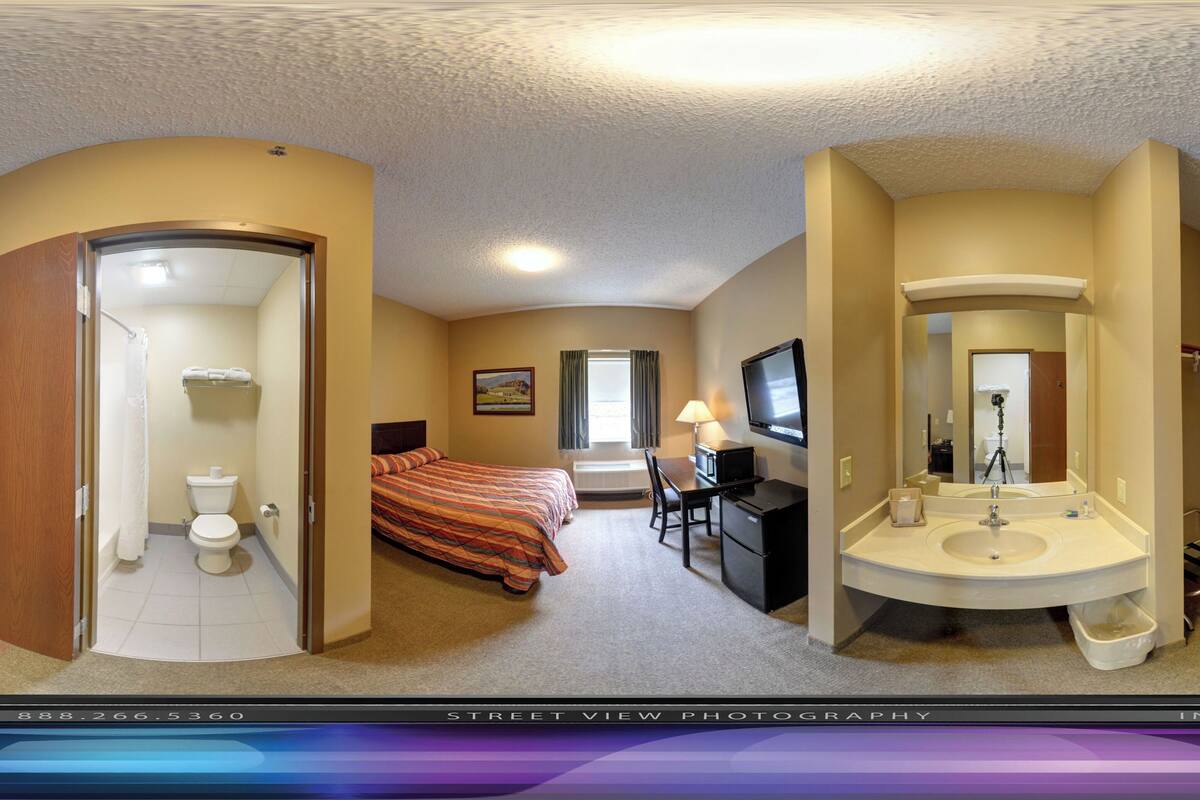 Una camera d'albergo con un letto, una scrivania, un bagno con lavandino e specchio, e una porta.