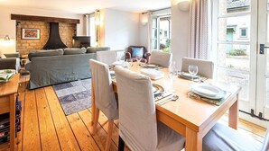 A cozy living room with a fireplace, a dining area with a table set for two, and a view of the outside through a large window. - Mill Village 59, Millstream Cottage (P) - On-site Facilities, Pool, Sleeps 6 (Somerford Keynes)