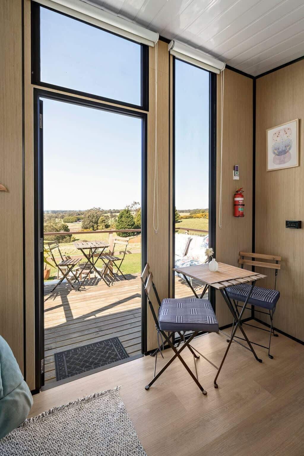 Une pièce dotée de grandes fenêtres, d’une terrasse en bois, d’une table avec des chaises et d’un extincteur fixé au mur.