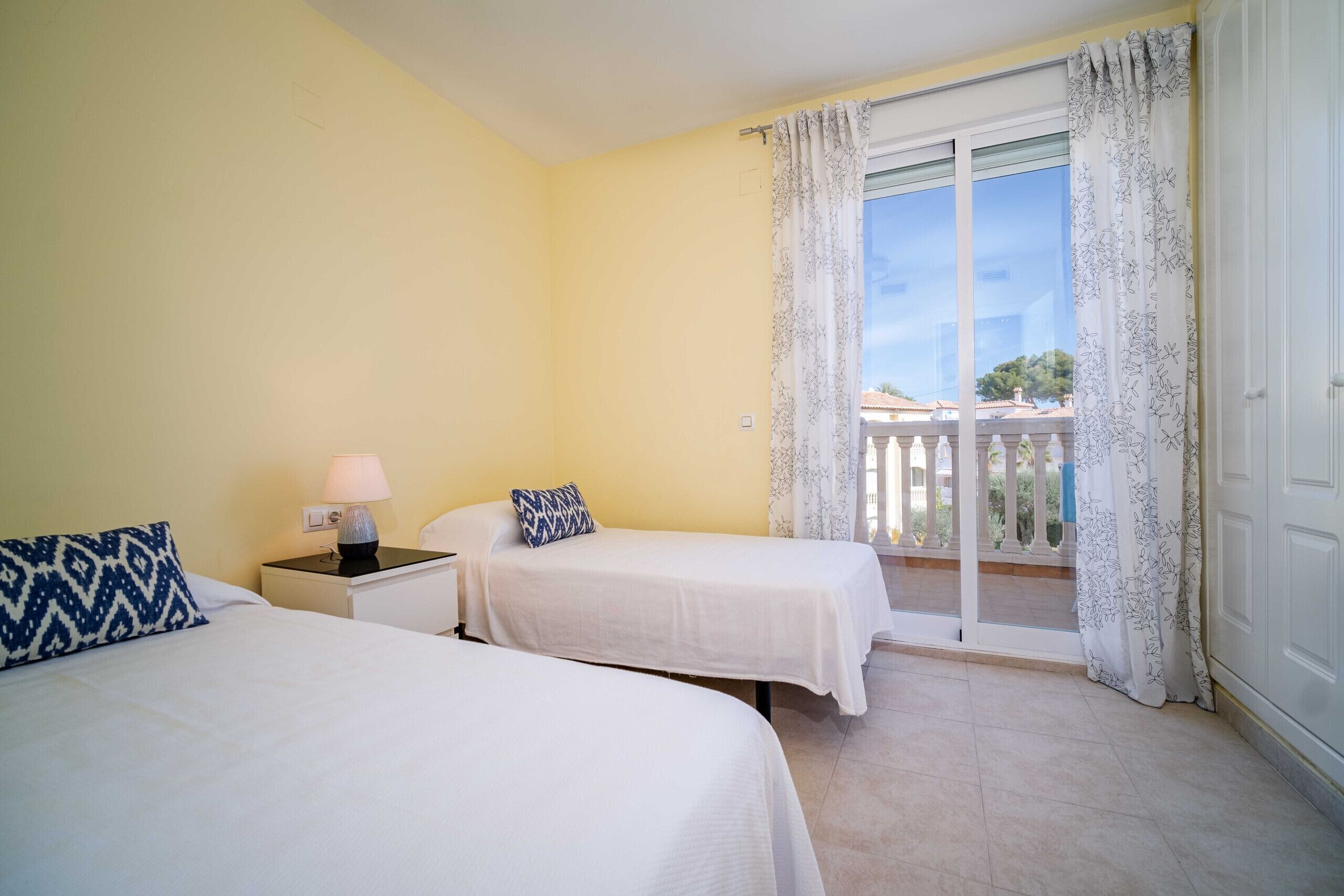 A hotel room with two beds, a nightstand with a lamp, and a balcony with curtains.