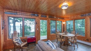A cabin with wooden walls and large windows, a dining area with a table and chairs, and a red cooler on the floor. - Lake Superior Cabin, 4BR, Sleeps 10, Private Beach (Paradise)