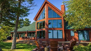 A two-story wooden cabin with large windows, outdoor seating, and a covered patio area. - Lake Superior Cabin, 4BR, Sleeps 10, Private Beach (Paradise)