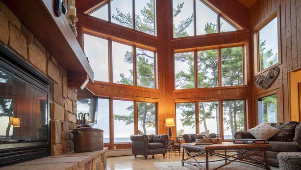A spacious living room with large windows, a stone fireplace, and wooden beams. - Lake Superior Cabin, 4BR, Sleeps 10, Private Beach (Paradise)