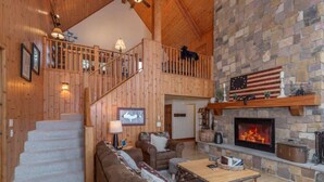 A spacious living area with a stone fireplace, wooden staircase, and a dog on the balcony. - Lake Superior Cabin, 4BR, Sleeps 10, Private Beach (Paradise)