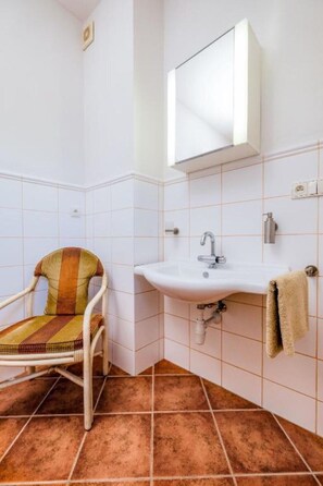 Ein Badezimmer mit weißen Fliesen, einem weißen Waschbecken, einem Spiegel über dem Waschbecken, einem Stuhl mit gestreifter Sitzkissen und einem Handtuch an der Wand.