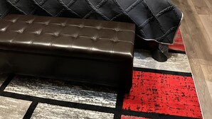 A bedroom with a bed, two nightstands, a lamp, a bedside table, a bench, and a red and white rug.