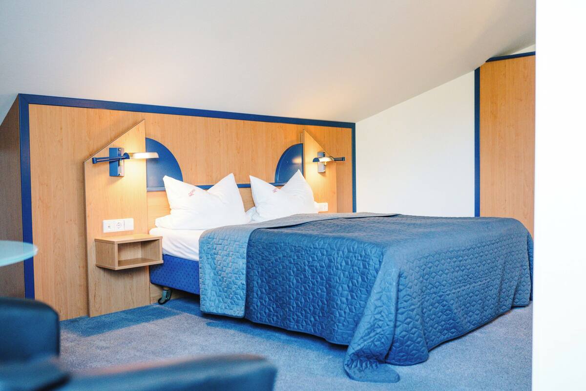 A neatly made bed with a blue quilt, a wooden headboard with built-in lighting, and a small bedside table with a lamp.