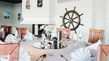 A dining table set with white tablecloths, silverware, and wine glasses, with a ship's wheel decoration on the wall.