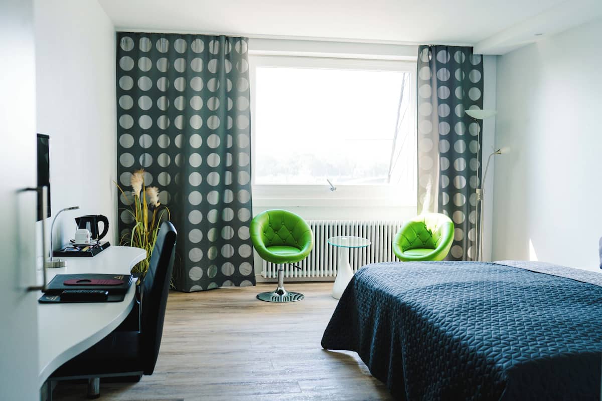A hotel room with a bed, two chairs, a small table, a radiator, and a window with curtains.
