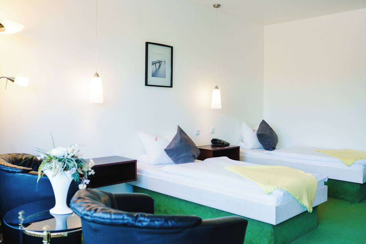 A hotel room with two single beds, a green carpet, a white vase with flowers, a blue armchair, and a desk with a lamp.