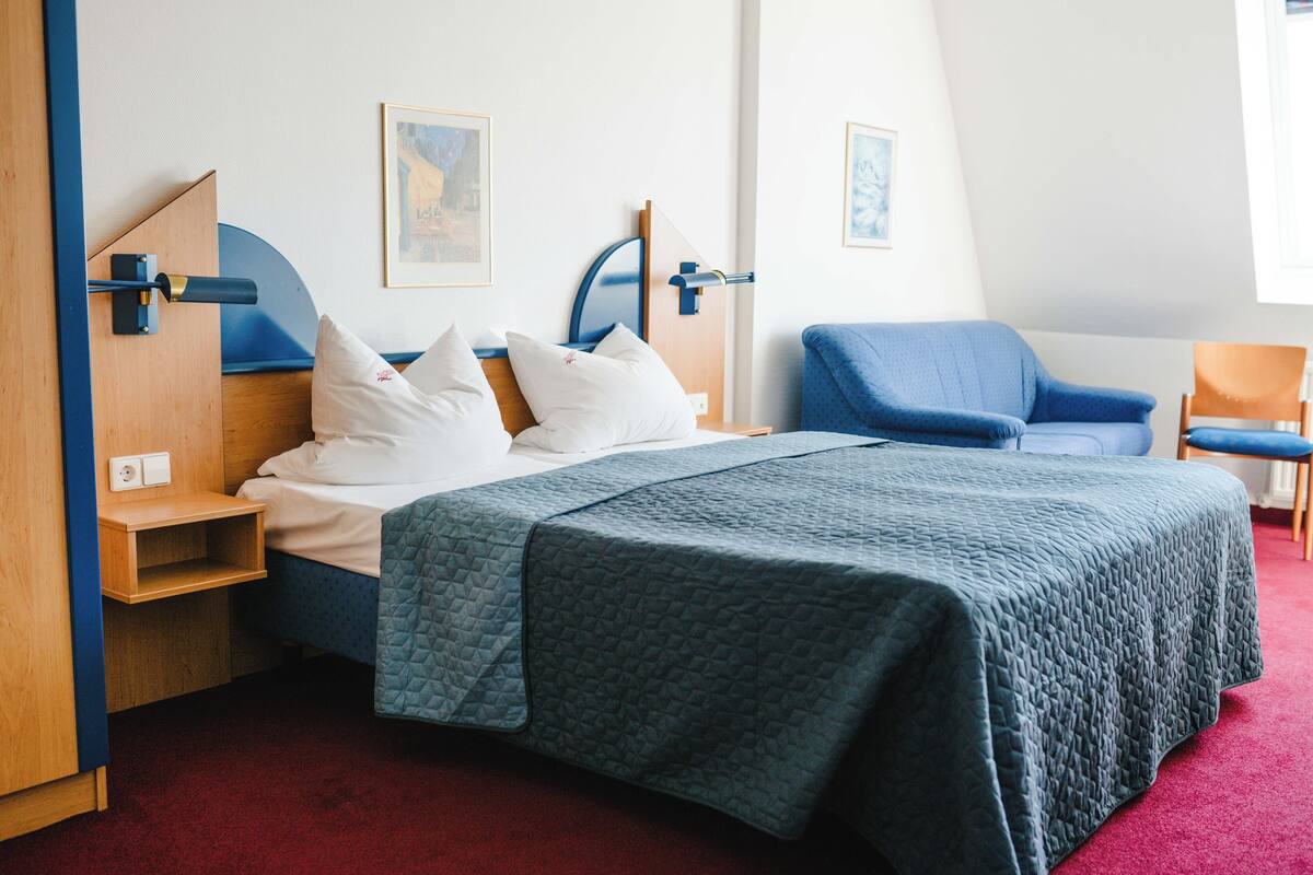A bedroom with a bed, a blue chair, a wooden nightstand, and a red carpet.