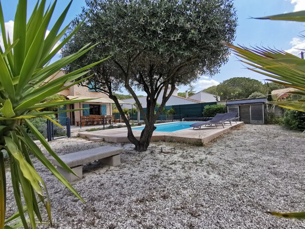 Zona de piscina con un árbol, una zona de estar cubierta y un edificio al fondo.
