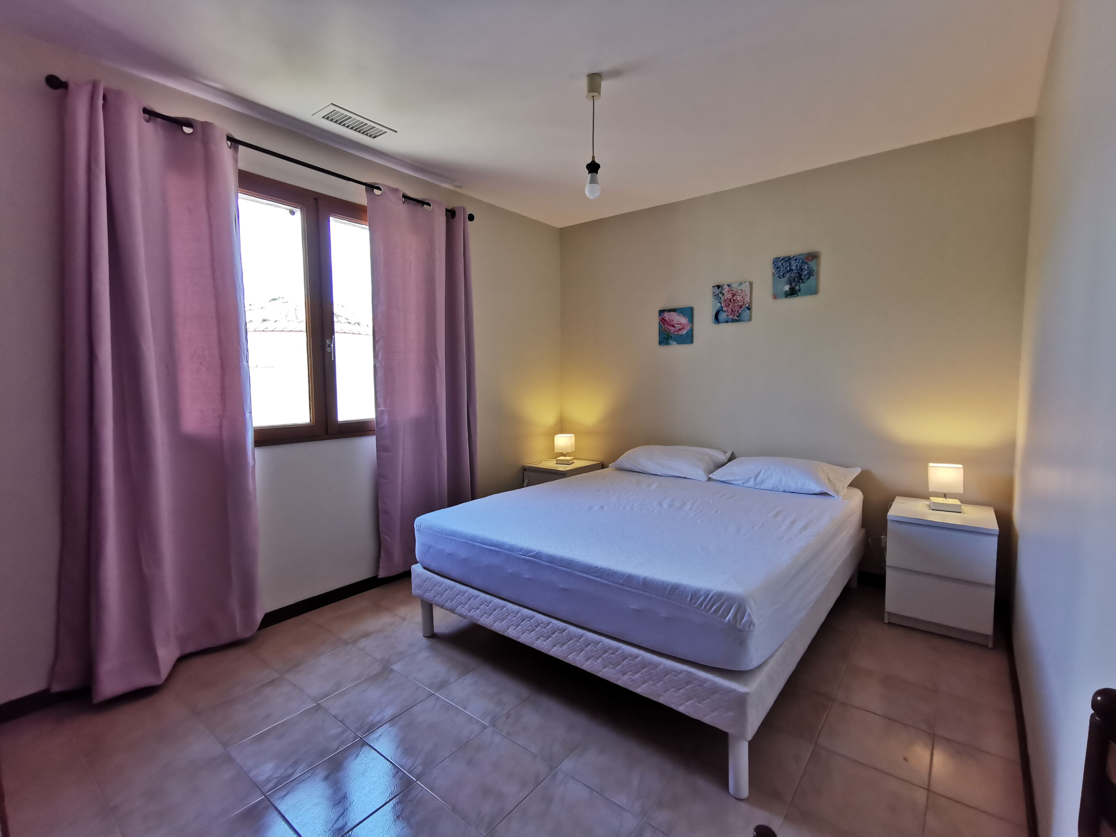 Un dormitorio con una cama, dos mesitas de noche con lámparas, una ventana con cortinas y una pared decorada con dos cuadros.