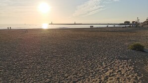Ein Strand mit einem Leuchtturm in der Ferne, Menschen, die am Ufer entlanggehen, und einem klaren Himmel.