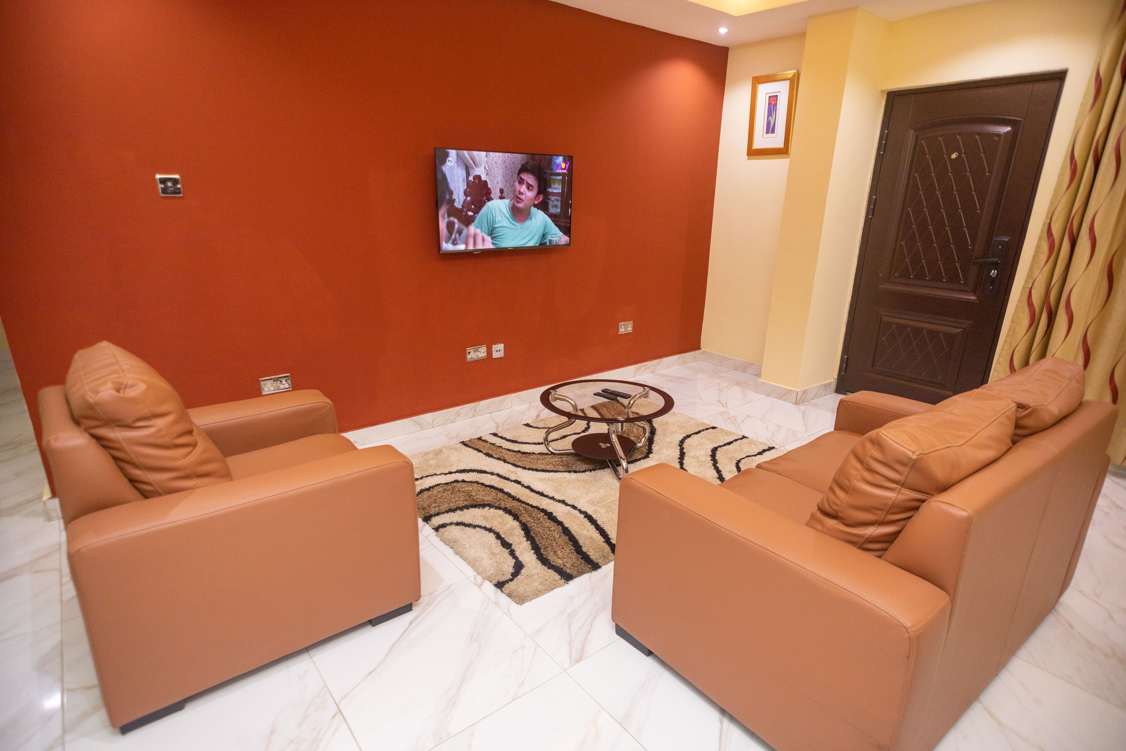 Una sala de estar con dos sofás de cuero marrón, una mesa de centro, un televisor montado en la pared, un tapete estampado y una puerta con un diseño decorativo.