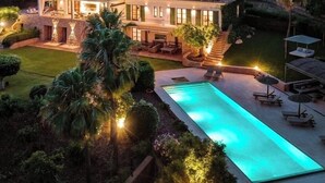 Une grande maison à deux étages, dotée d’une piscine, entourée d’une végétation luxuriante et avec des collines en arrière-plan.