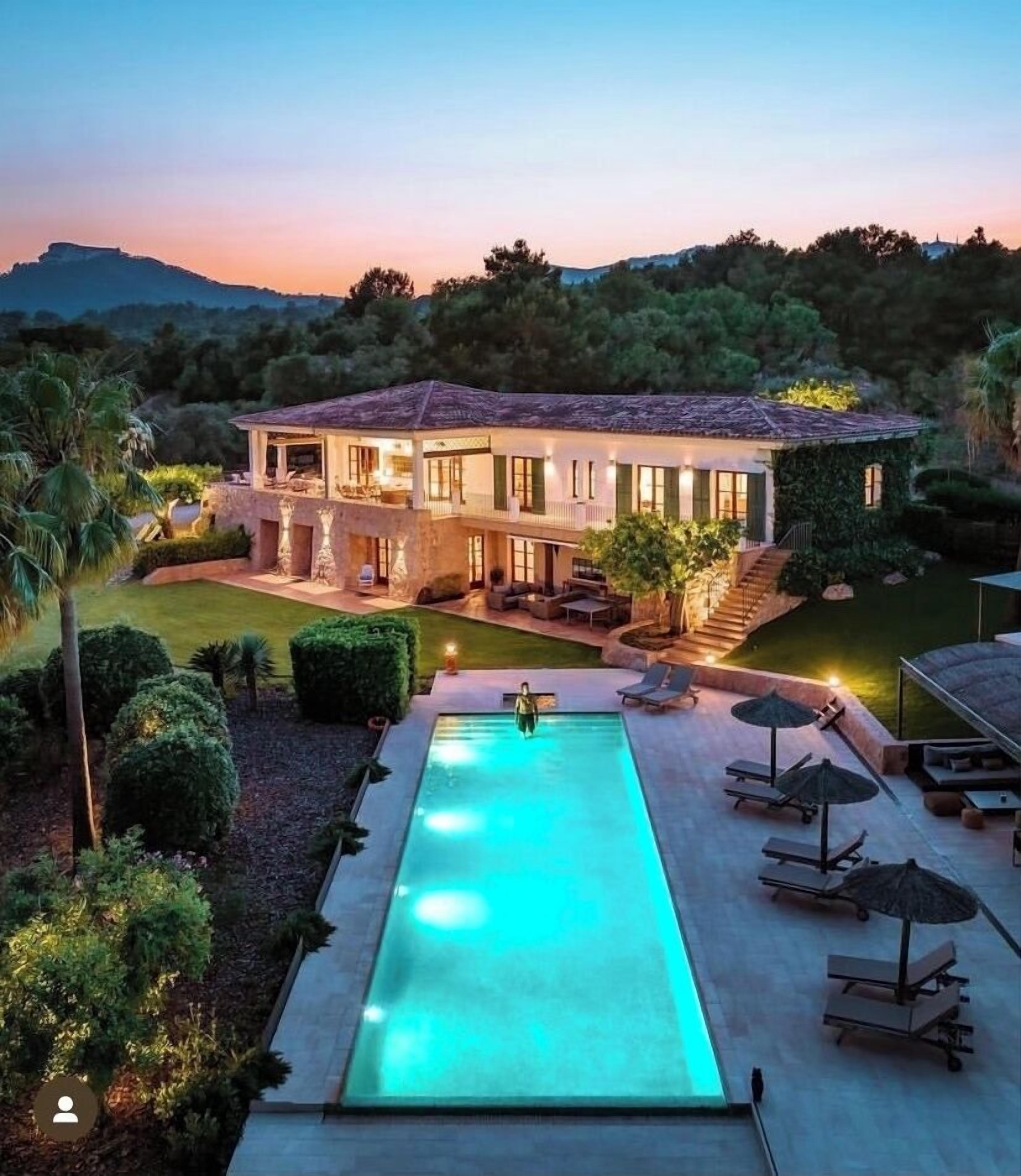 A large, two-story house with a swimming pool, surrounded by lush greenery and hills in the background.