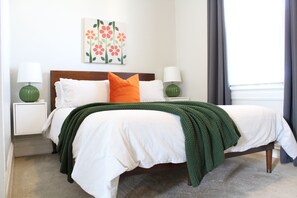 Een opgemaakt bed met wit beddengoed en een groene deken, met aan beide zijden nachtkastjes met lampen, in een kamer met een effen muur en een raam met gordijnen.