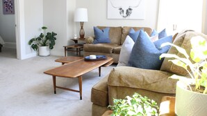 Una sala de estar con sofá color beige, cojines azules, una mesa de centro de madera y una planta en maceta.