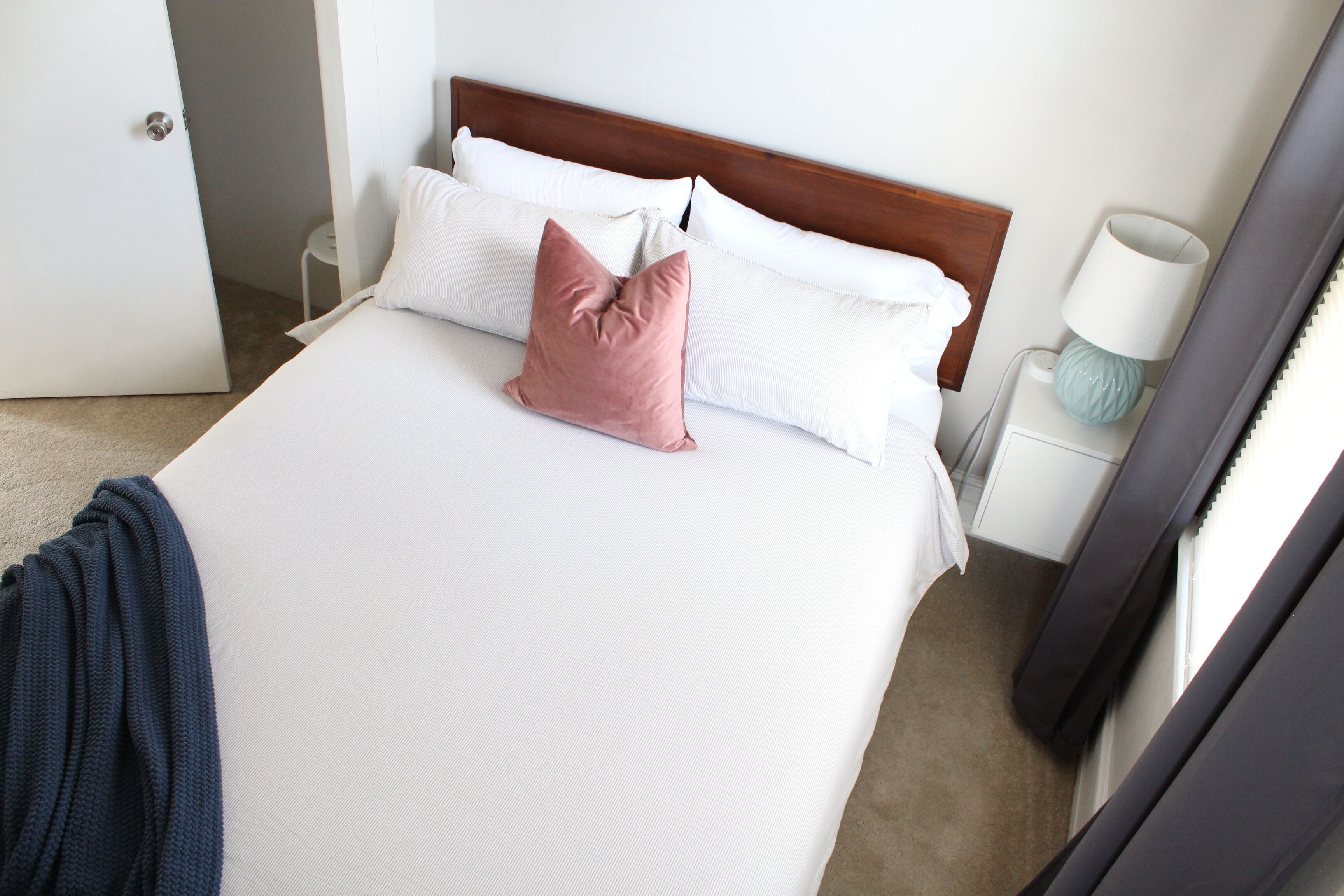 A neatly made bed with white and pink pillows, a wooden headboard, a bedside lamp, and a small white nightstand.