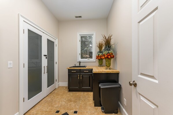 A narrow hallway with a kitchen area featuring dark cabinets, a sink, and a trash bin. There is a window with a view of trees and a decorative plant arrangement on the counter. - Abbey Manor on Pinehurst #6- Golf Views -Sleeps 24 (Pinehurst)