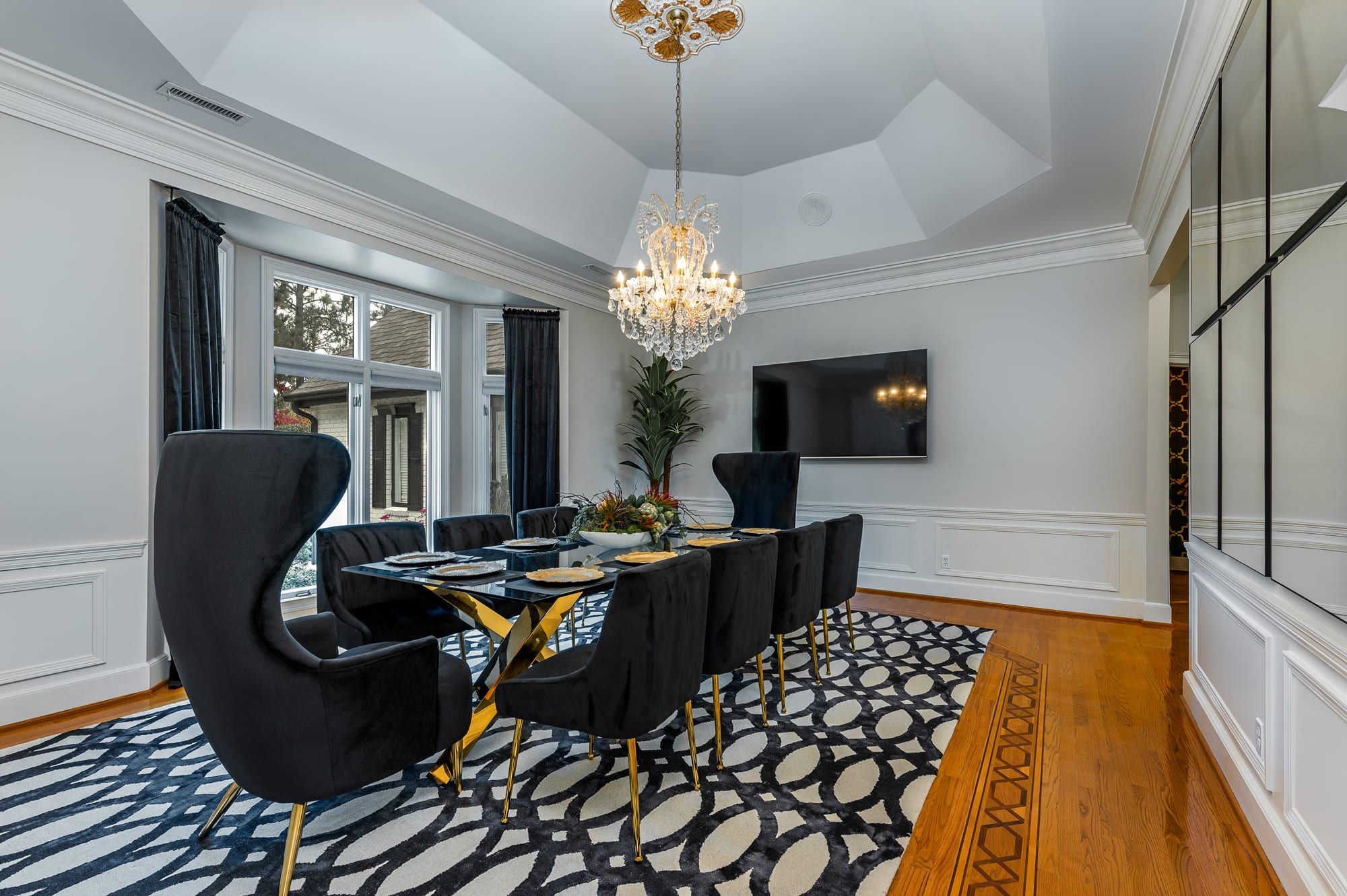 A dining area with a long table, black chairs, a chandelier, and a patterned rug.